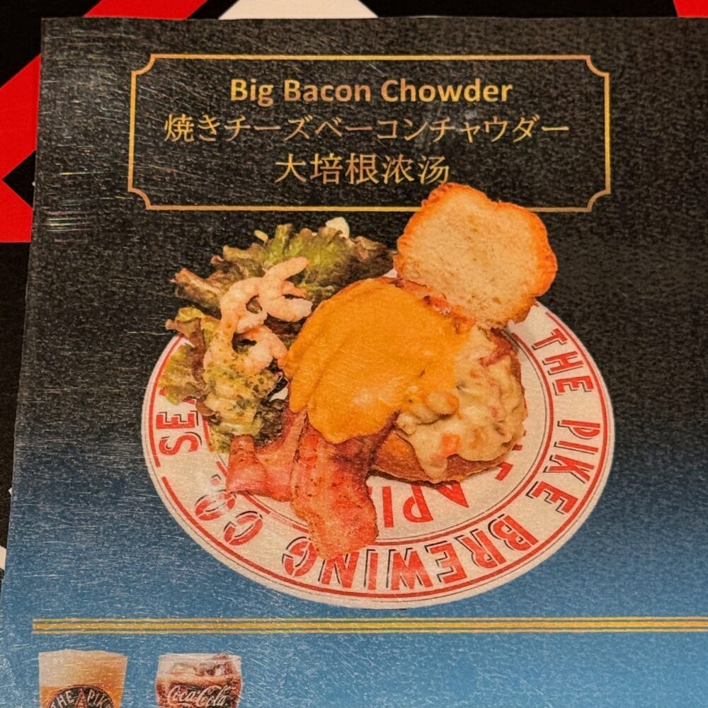 プライオリティパス専用セットメニュー「焼きチーズベーコンチャウダー(Big Bacon Chowder)」の案内カード。えびのフリットや野菜を添えたチャウダーが白いプレートに盛られた写真と、選べるドリンクの説明が記載されている。