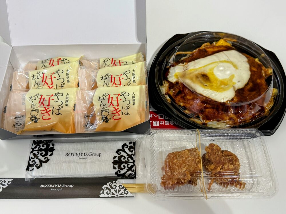 Japan Traveling Restaurant by BOTEJYU（ぼてぢゅう）のテイクアウトセットの俯瞰写真。右側に目玉焼きが乗ったお好み焼きの容器、手前に唐揚げ2個入りの容器、左側の箱に個包装された「なんば好っきや」の大阪土産（お菓子）が6個入っている。下に「BOTEJYU Group」と書かれた箸袋と紙ナプキンがある。