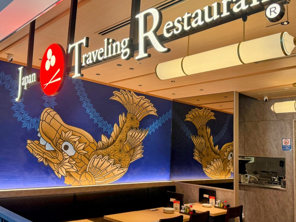 Japan Traveling Restaurant by BOTEJYUの店内の壁面。青い背景に、金色で装飾された大きな鯱（しゃちほこ）のような生き物のイラストが描かれている。上部には「Japan Traveling Restaurant」の看板、手前には木目調のテーブルと黒いソファ席が見える。