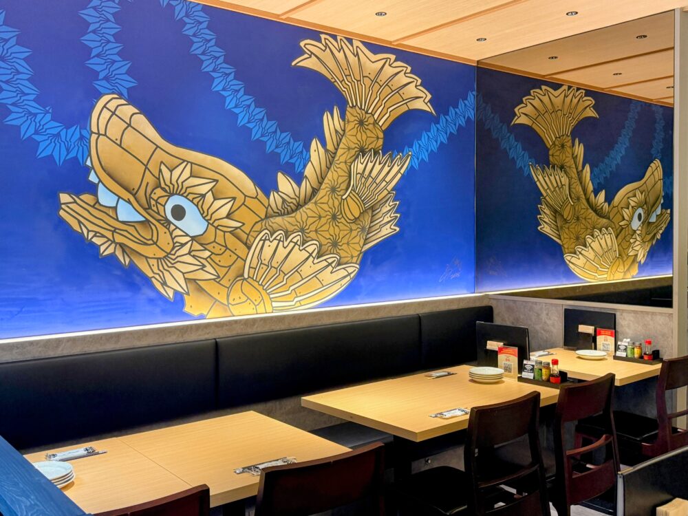 Japan Traveling Restaurant by BOTEJYUの店内風景。壁には金色の迫力ある鯱の壁画が描かれ、モダンな雰囲気のテーブル席が並んでいる。