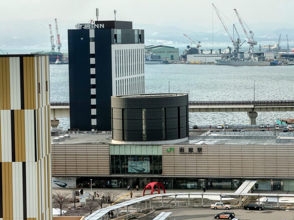 フォーポイントフレックス函館の客室から見下ろすJR函館駅の全景と、奥に広がる函館港の景色。