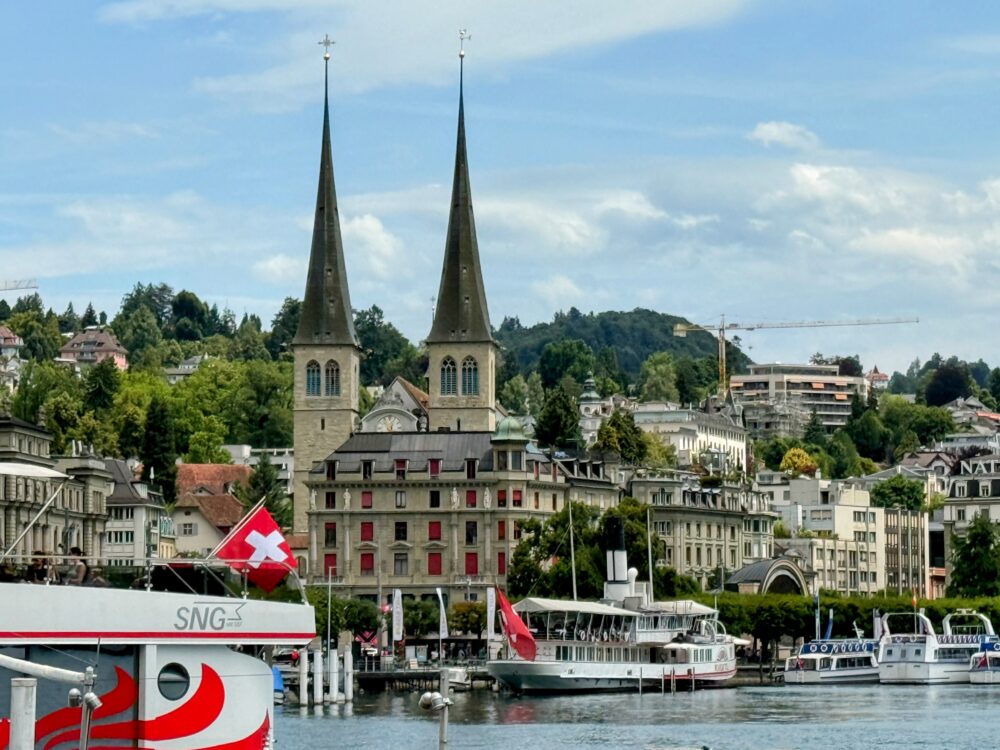 ルツェルン湖畔から望むホーフ教会の双塔。スイス国旗を掲げた船と湖畔の街並みが広がる風景