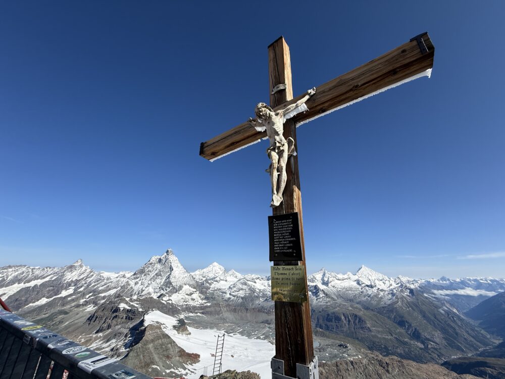 マッターホルン・グレッシャーパラダイスの展望台からの大パノラマ。雪に覆われた山々が広がり、頂上には白いキリスト像の十字架が立つ。眼下には氷河と遠くの山々が見渡せる。