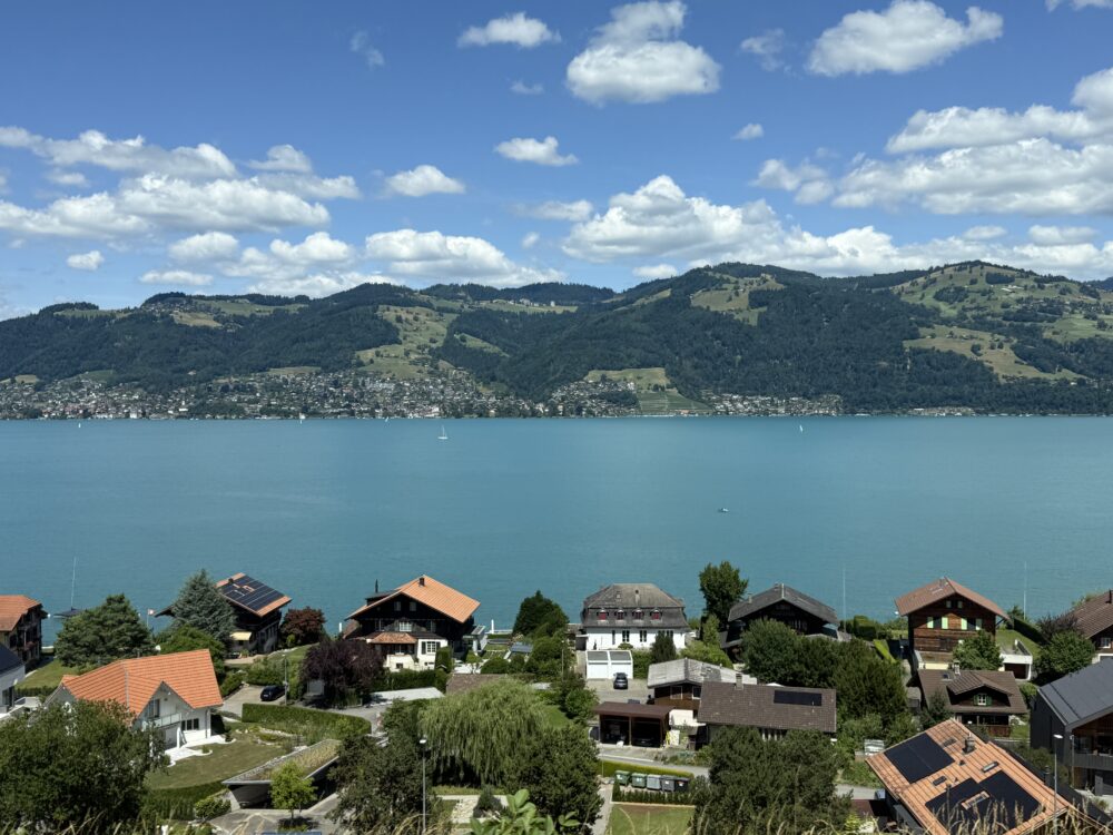 ツェルマットからチューリッヒへ向かうSBB鉄道の車窓から望む湖畔の風景。青く澄んだ湖と緑の山々、赤い屋根の家々が並ぶスイスらしい美しい田園風景が広がる。