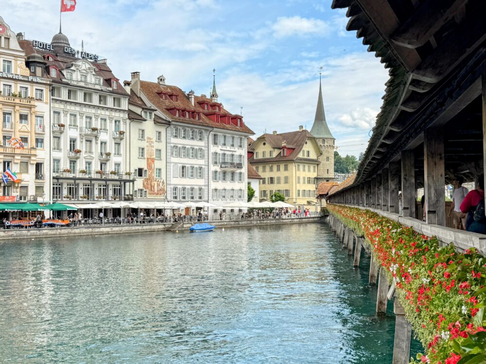 ルツェルンのカペル橋から見たロイス川沿いの旧市街。川沿いにはカラフルな古い建物が並び、その手前には花で飾られたカペル橋の側面が見える。遠景には尖塔が見える。