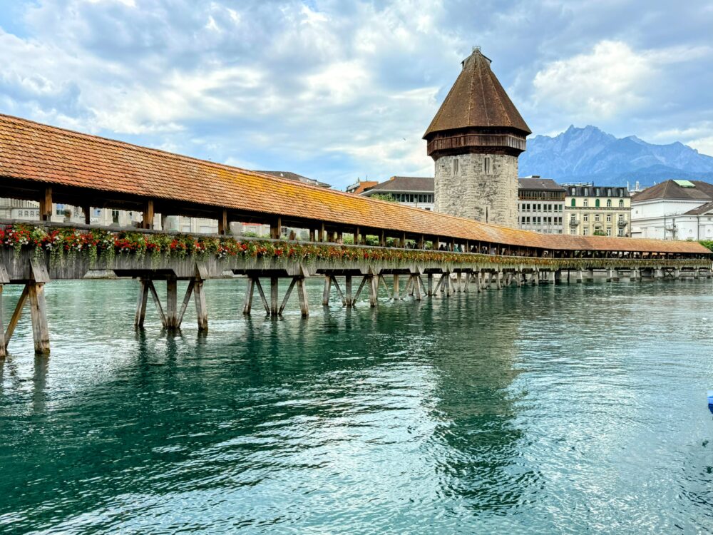 スイス・ルツェルンの象徴カペル橋。赤いペチュニアの花が欄干を彩り、背後には水の塔と青空が広がる美しい風景