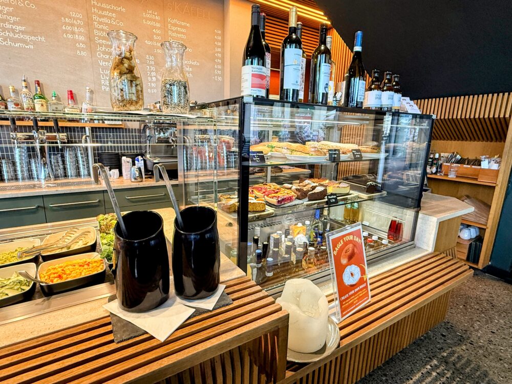 カフェのフードとドリンクの陳列ケース。様々な種類のタルトやケーキが並び、カウンターの上にはワインボトルが飾られている。