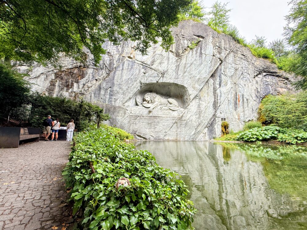 ルツェルンの瀕死のライオン像の全景。岩壁のレリーフが、手前の池のような水面と、周囲の木々の豊かな緑に囲まれている。左側には観光客の姿が見える。