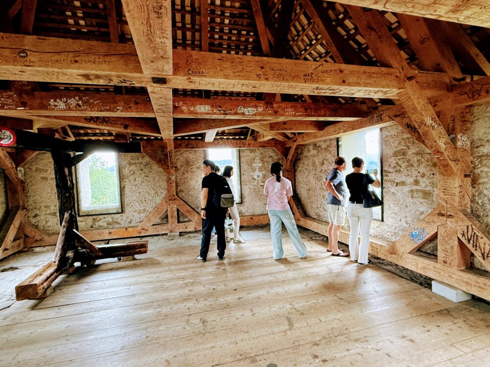 ムーゼック城壁の塔の内部。木造の骨組みがむき出しの屋根裏部屋に、窓から外の景色を眺める数人の観光客が立っている。