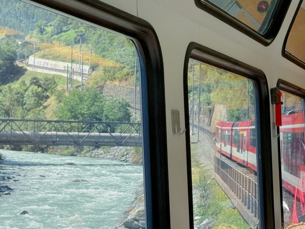 スイスの鉄道の車窓から見た景色。手前には勢いよく流れる川と鉄橋があり、窓の右側にはカーブを曲がる自分の乗る赤い列車の車両の一部が見えています。背景には緑豊かな山肌が広がっています。