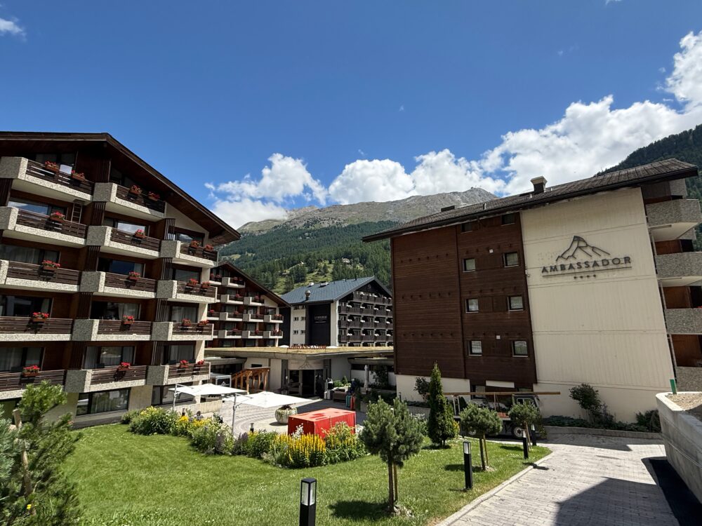 青空の下に建つホテル アンバサダー ツェルマットの外観。山小屋風の建物が並び、バルコニーには赤いゼラニウムの花が飾られている。背景にはツェルマットの山々がそびえ、リゾートらしい雰囲気が漂っている。