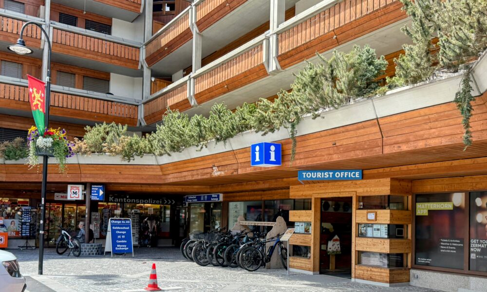 スイスのツェルマット駅前にある観光案内所の外観。木製の温かみのある建物で、青いインフォメーションマークと『TOURIST OFFICE』の文字が見えます。建物の手前には自転車が数台停められ、左側には小さなショップや警察署の看板も見えます。上階はバルコニー付きの宿泊施設のようです。晴れた日の日差しの中、観光客が行き交う賑やかな様子がうかがえます。」