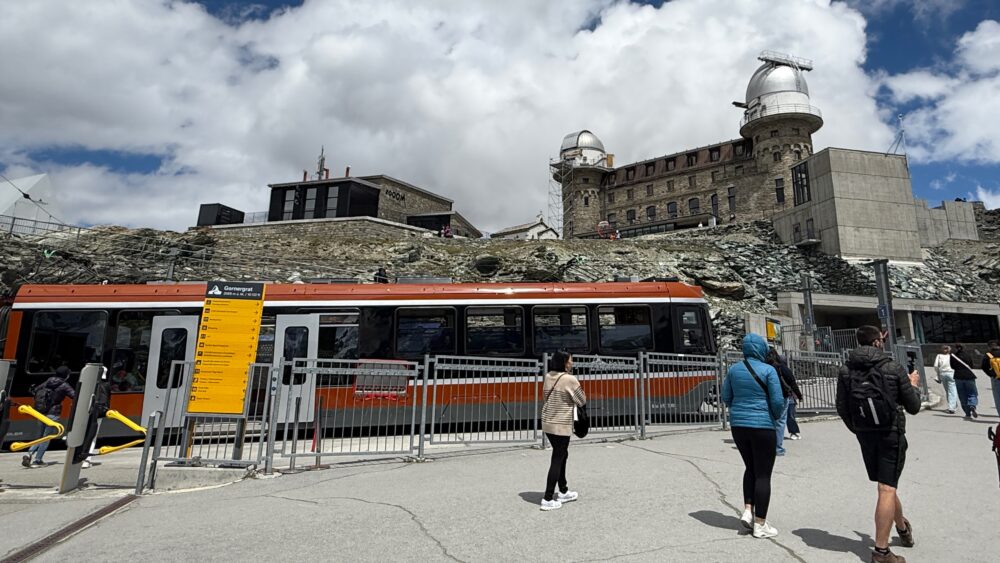 ゴルナグラート鉄道の終点駅ゴルナグラート駅に停車するオレンジ色の登山列車。背後にはツェルマットの名所・クルムホテル ゴルナグラートと天文台がそびえ、観光客が周辺を散策している様子。