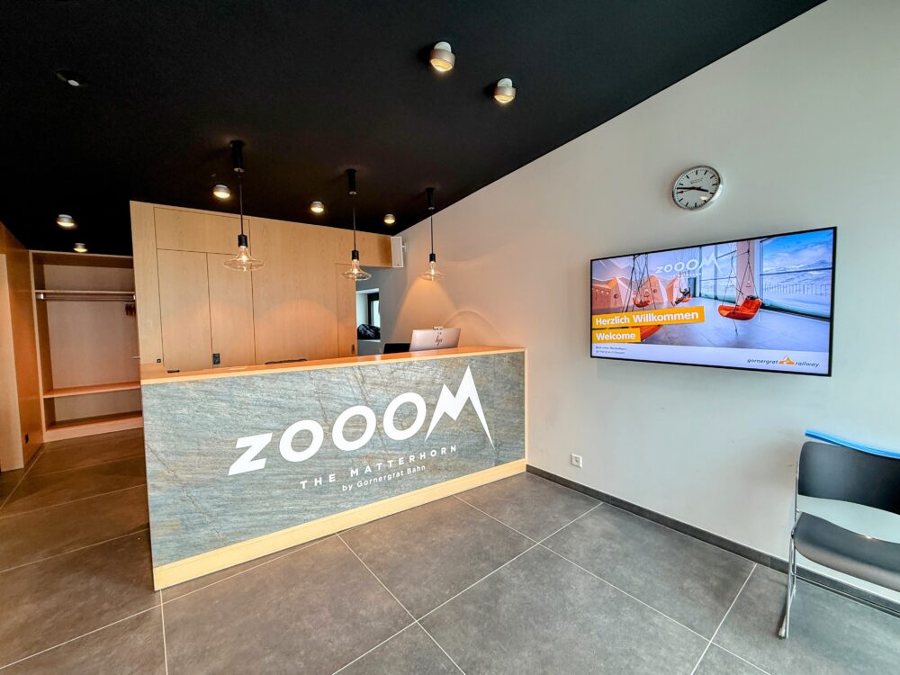 ゴルナーグラート展望台の施設内にある「ZOOOM THE MATTERHORN」の受付カウンター。モダンな木目調とグレーのタイルで統一された空間に、ブランドロゴとデジタルサイネージが設置されている。

