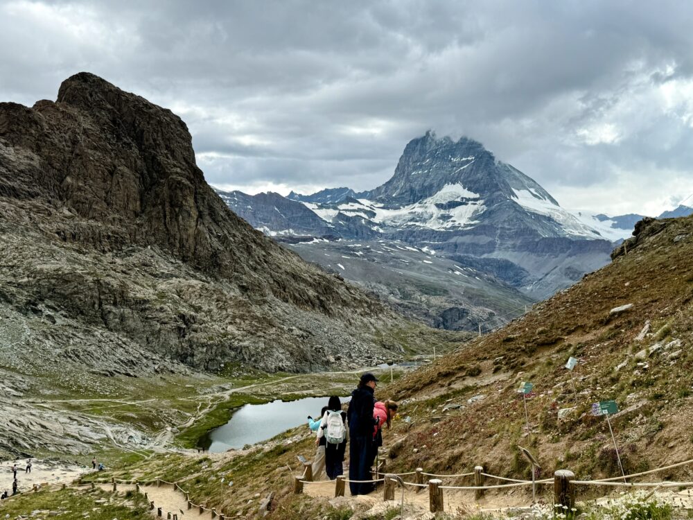 ハイキングコースを歩く人々。マッターホルンへ続く荒涼とした山肌のハイキング道と、遠くに湖（リッフェル湖）が見える。