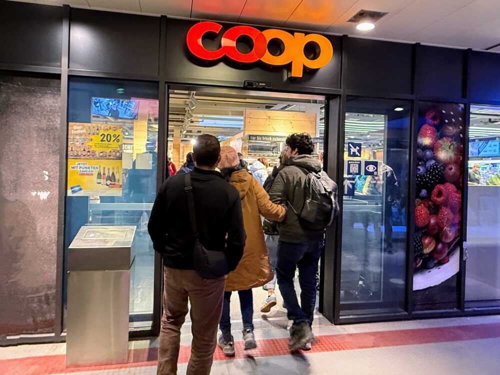 ツェルマットにあるスーパーマーケット「COOP（コープ）」の入り口。オレンジ色のロゴが目立ち、数人の客が店内に入っていく様子。