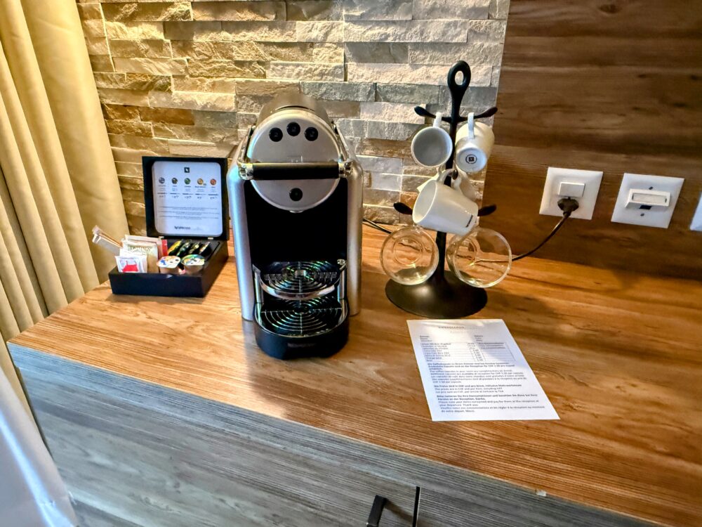 ホテルの客室に備え付けられたネスプレッソ（Nespresso）のコーヒーメーカーと、カプセル、カップ。木目調のカウンターに置かれており、後ろの壁は石目調のタイルになっている。