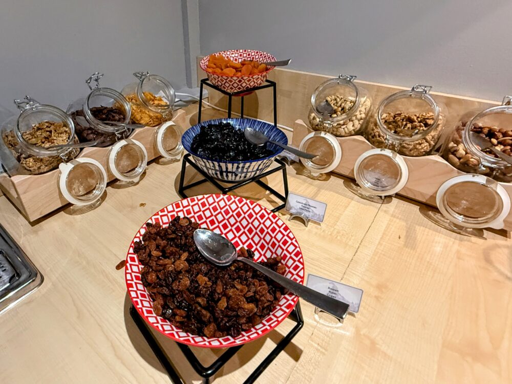 朝食ビュッフェのミューズリーバー。テーブルの上に、アーモンド、クルミ、レーズン、アプリコットなどのナッツやドライフルーツが、ガラス瓶やボウルに陳列されている。