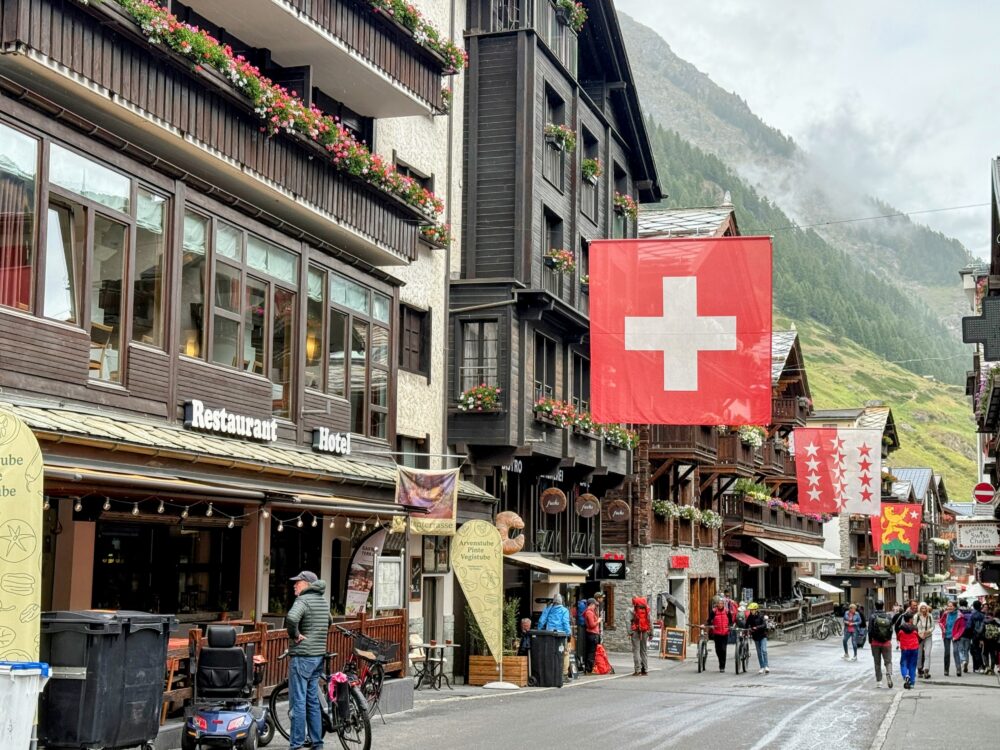 スイス国旗や州旗が飾られたツェルマット村の街並み。木と石造りの伝統的なシャレー建築が並び、窓辺には色とりどりの花が飾られている。