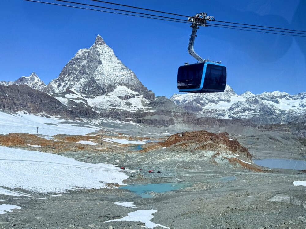 ロープウェイのゴンドラから見下ろす、雪と岩肌が広がる山岳地帯の景色。遠景にはマッターホルンがそびえ、眼下には氷河湖と赤いヘリコプターが小さく見える。