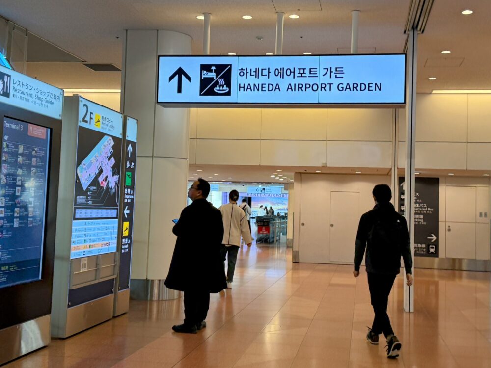 羽田空港第3ターミナル内の案内表示。ホテルと羽田エアポートガーデンへの方向を指しており、人々が連絡通路に向かって歩いている。