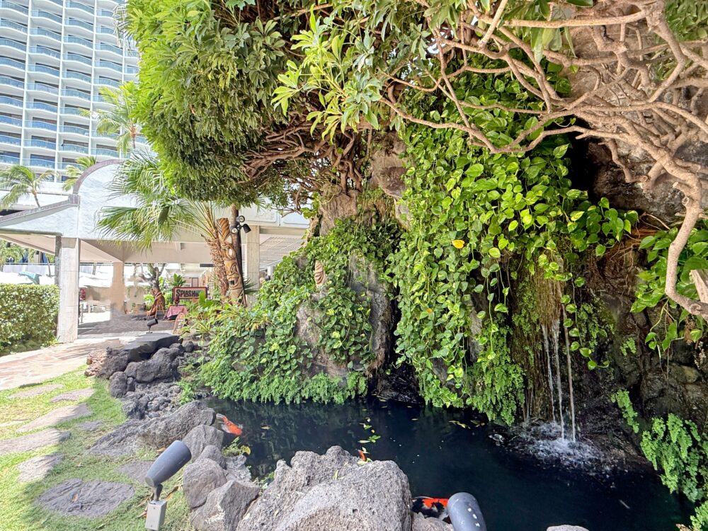 ホテル中庭の一角にある滝と池の風景。緑のつる植物が茂る岩壁から水が流れ落ち、池の中では色鮮やかな鯉が泳いでいる。