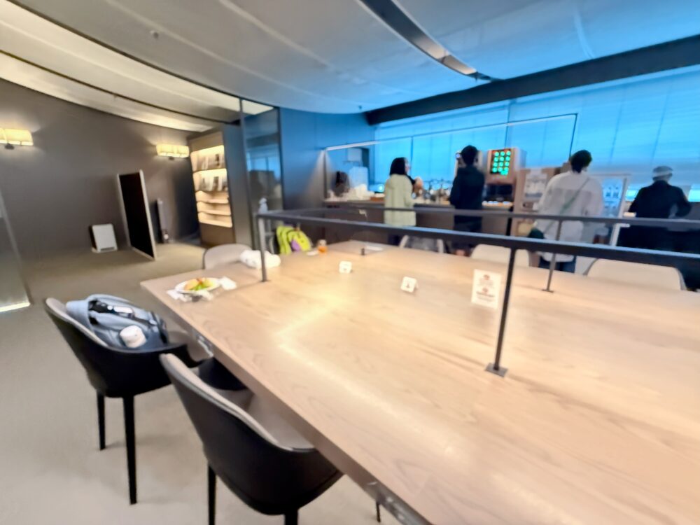 ラウンジ中央の大テーブル席。背面にはドリンクカウンターがあり、多くの利用者が軽食を楽しんでいる様子。