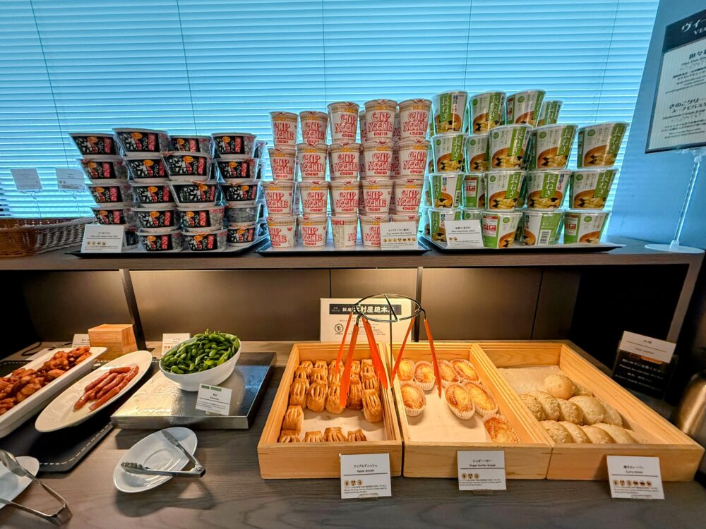 ラウンジの軽食コーナー。パン、枝豆、ソーセージ、唐揚げ、焼きそば、カップヌードルなどが並ぶビュッフェ台。