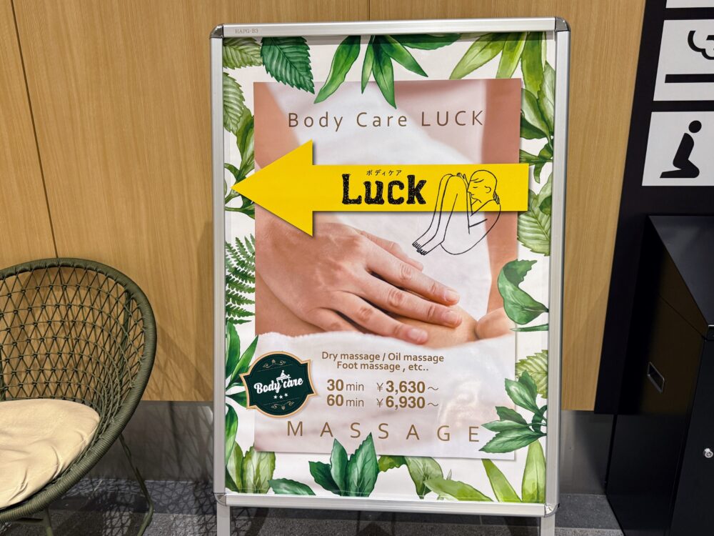 Body Care LUCKの看板。マッサージの種類（ドライ、オイル、フット）と30分3,630円からの料金が表示されており、店への方向を示す黄色の矢印が付いている。