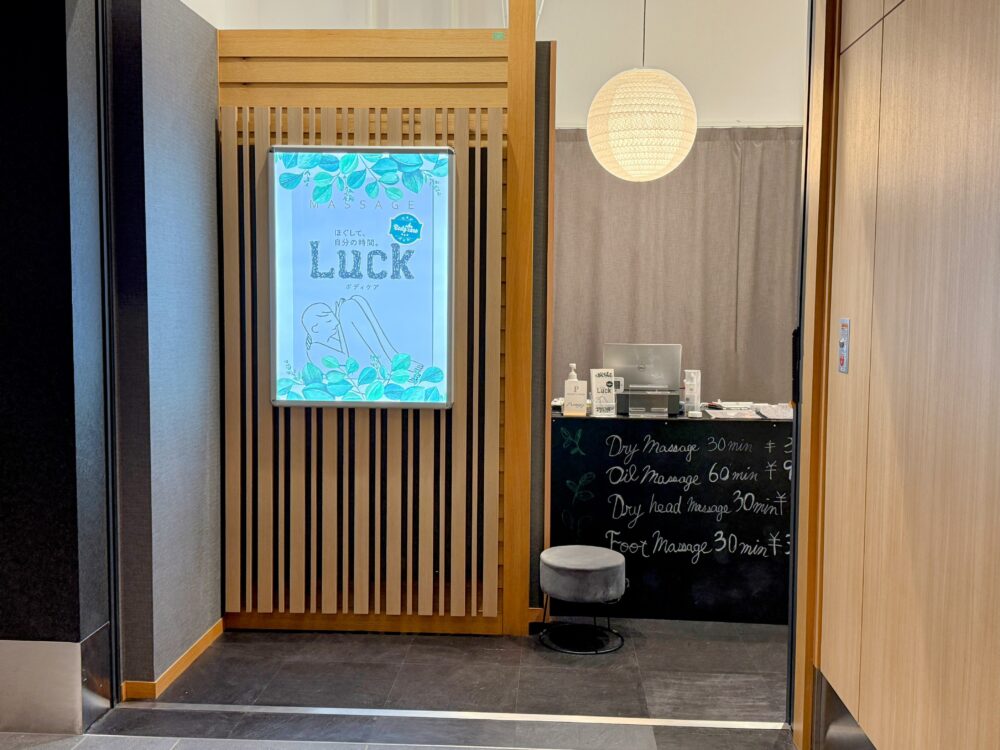 Body Care LUCKの受付の様子。木製のルーバーと丸いペンダントライトが特徴的で、黒板にドライマッサージやオイルマッサージなどの料金が手書きで表示されている。