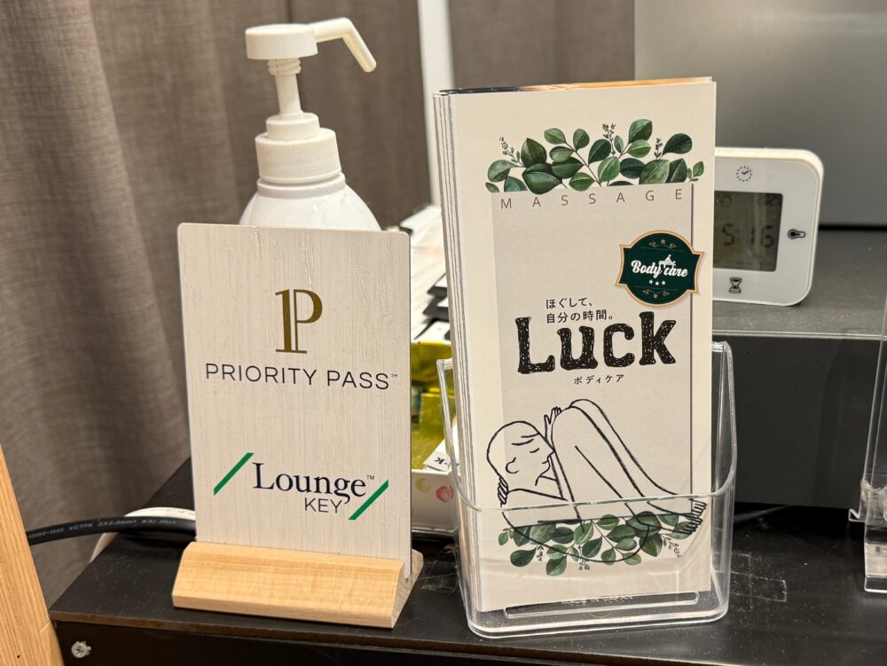 Body Care LUCKの受付にある、プライオリティ・パス（Priority Pass）とラウンジ・キー（LoungeKey）が利用できることを示す立て札。横には店舗のパンフレットが置かれている。