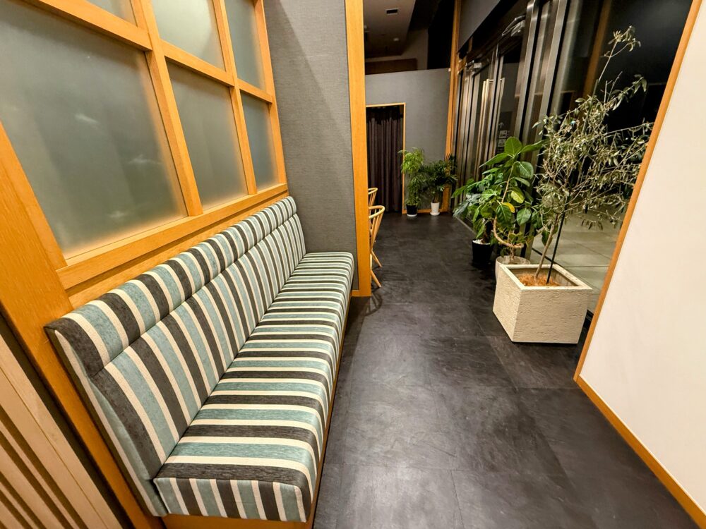 店内の待合スペースにあるストライプ柄の長いソファ（ベンチ）。木枠と観葉植物が配置され、リラックスできる空間。