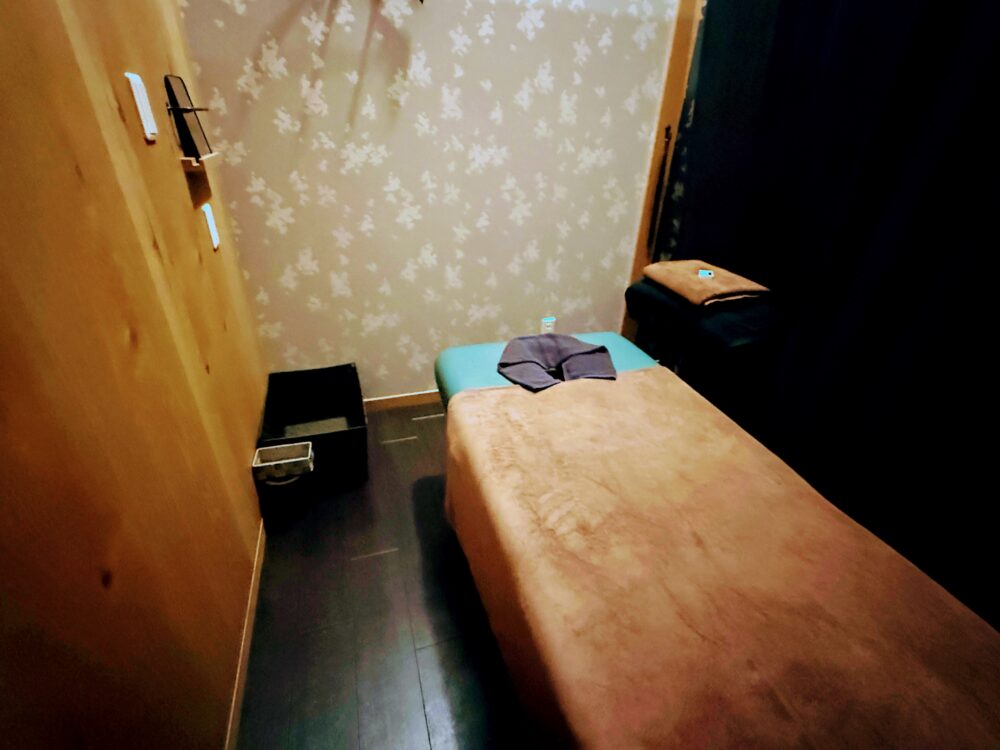 施術室の様子。落ち着いた照明で、タオルケットがかけられた施術用ベッドと、荷物を入れるカゴが置かれている。