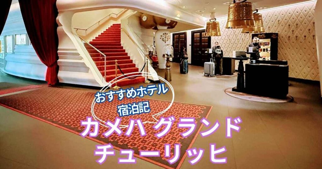 カメハ グランド チューリッヒのロビー。赤い絨毯の階段と金のベル型シャンデリアが印象的なデザイン空間。中央に「おすすめホテル 宿泊記 カメハ グランド チューリッヒ」という文字が入っている。