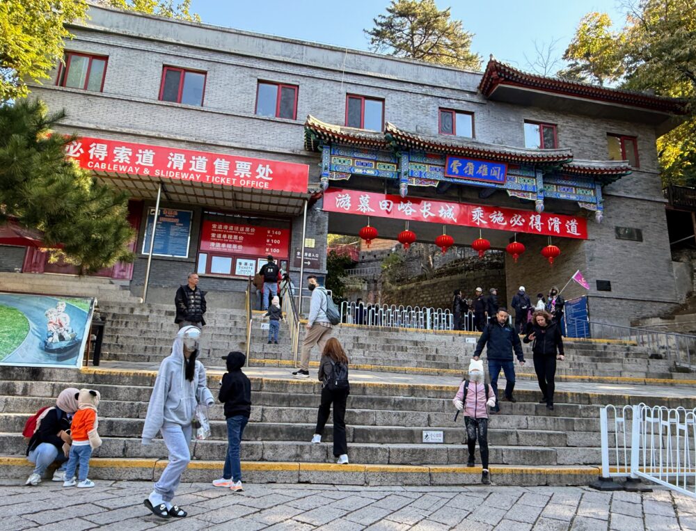 慕田峪長城リフト乗り場の入口。チケットオフィスと「CABLEWAY & SLIDEWAY TICKET OFFICE」の赤い看板があり、多くの観光客が行き交っている様子。