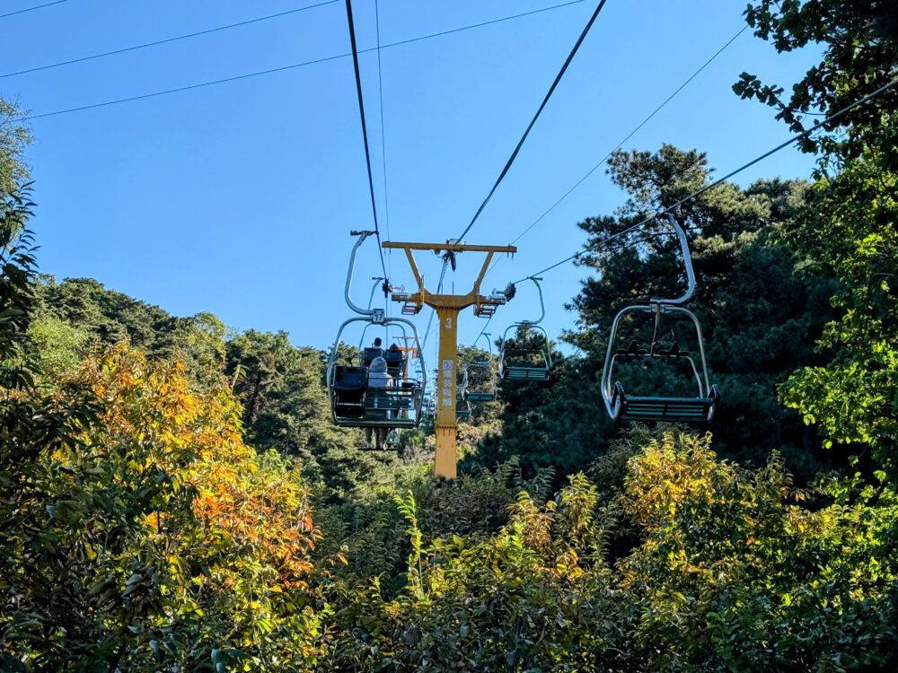 慕田峪長城のリフトに乗って山を登る途中の景色。緑に囲まれた中を進み、秋の木々と澄んだ青空が広がる。
