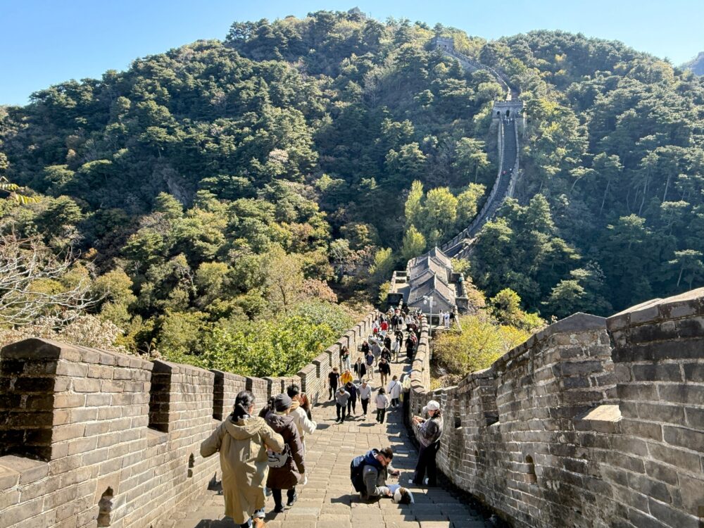 慕田峪長城の右方向（1号大角楼方面）へ向かう途中の坂道。
傾斜のある階段を多くの観光客が登っている様子。両側に緑が広がり、青空が美しい。