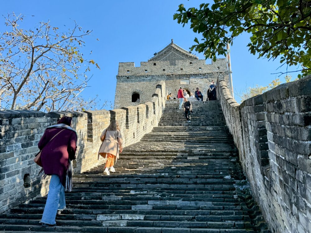 慕田峪長城の右方向（1号大角楼方面）へ向かう途中の坂道。
傾斜のある階段を多くの観光客が登っている様子。両側に緑が広がり、青空が美しい。