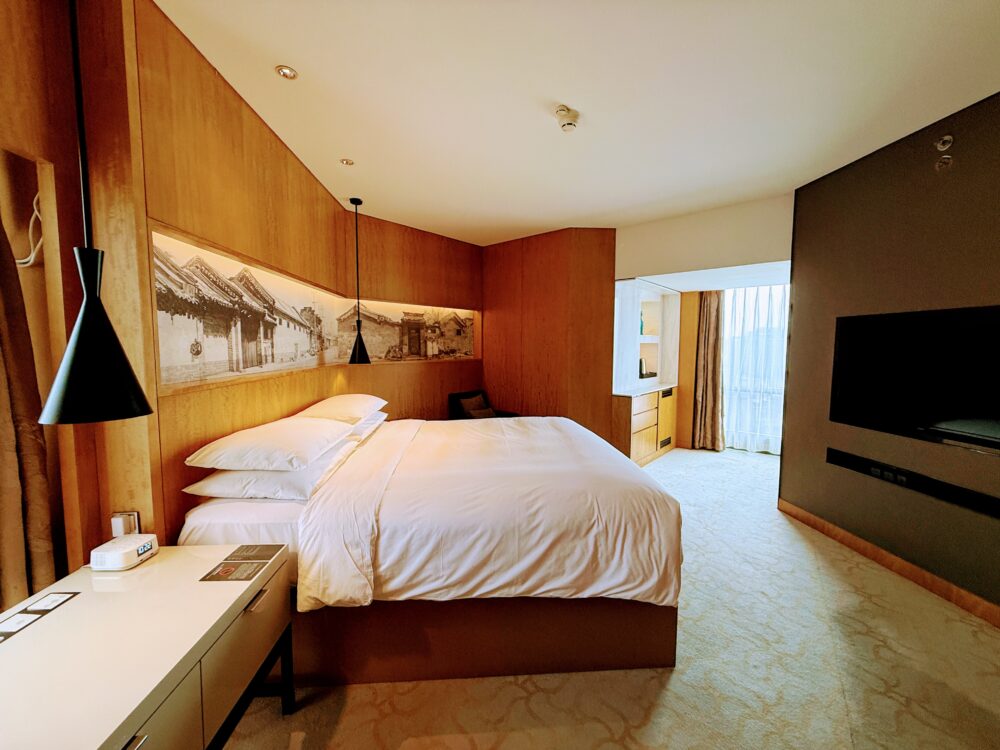 ルネッサンス北京王府井ホテルのスイートルーム。温かみのある木目調のインテリアで、ベッド背面には北京の胡同（フートン）を描いたアートが飾られている。広々とした空間と大きな窓が特徴。