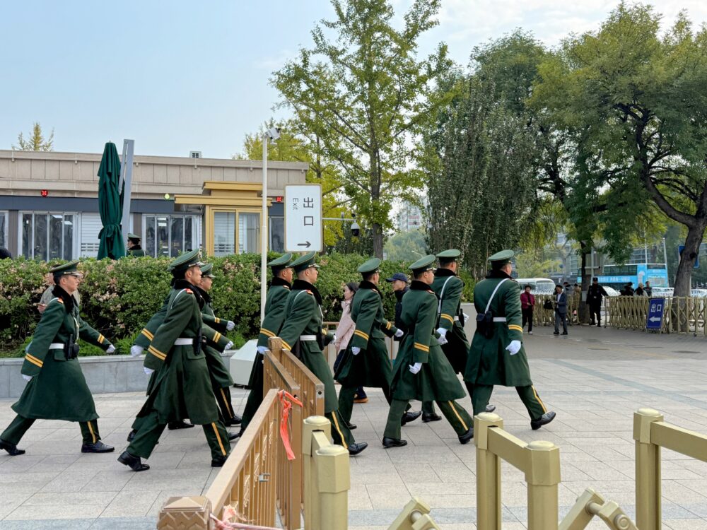 天安門広場の入口付近で、緑の制服を着た警備兵の隊列が歩いて移動している様子。広場の厳重な警備体制と観光地の緊張感が伝わる光景。