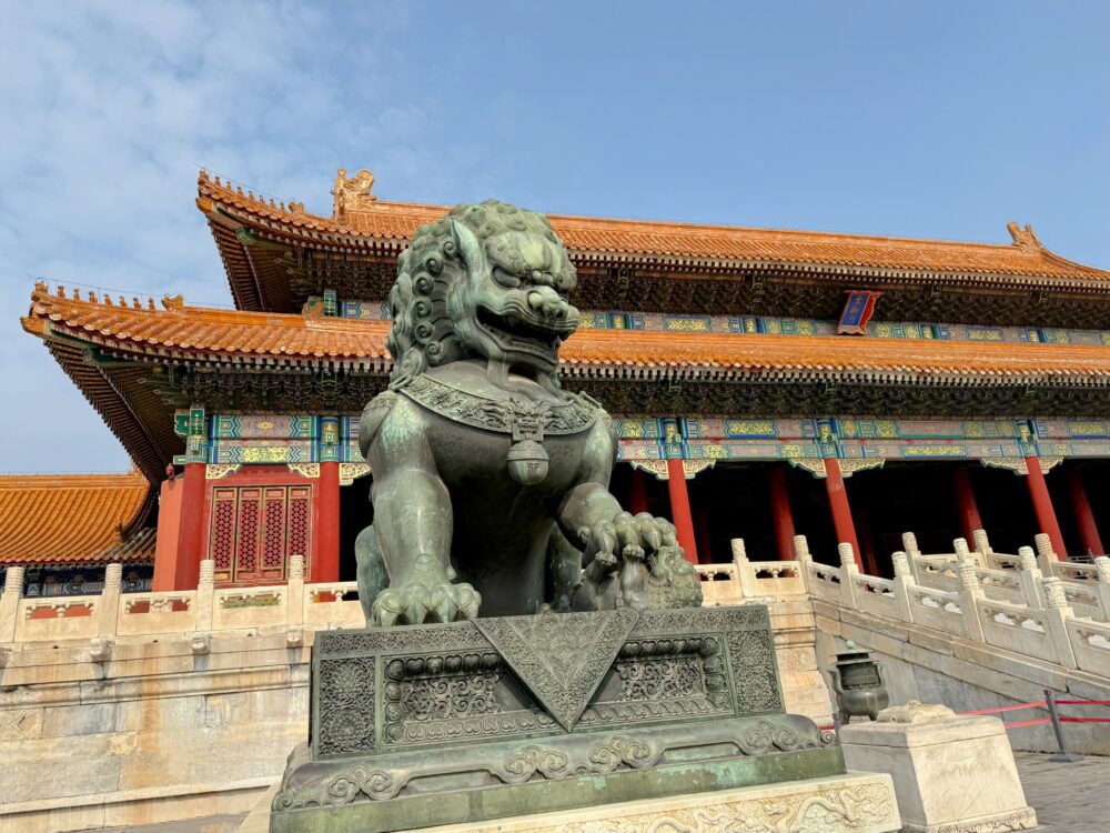 太和門の赤い柱と金瓦屋根を背景にした銅製の獅子像。力強い姿が強調されている。