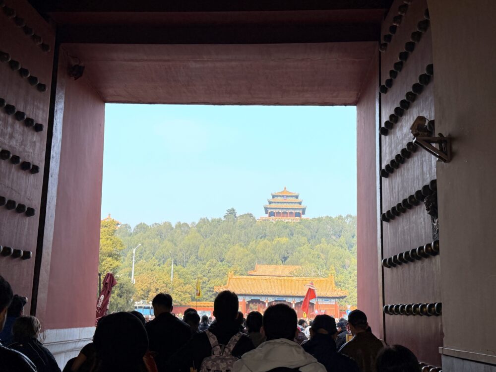 故宮・神武門の巨大な門の内側から外を眺め、正面に木々の丘と景山公園の万春亭が見えている写真。