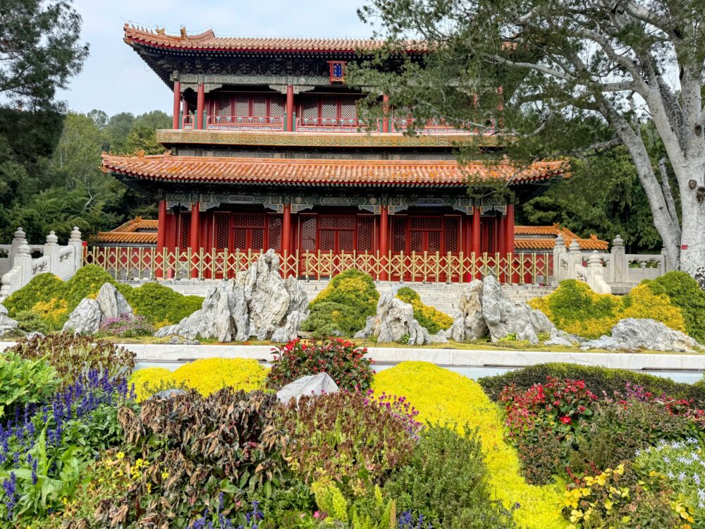 景山公園入口付近にある绮望楼を正面から撮影した写真。朱塗りの柱と黄瓦の二層建築が特徴で、前には色鮮やかな花壇と奇石が並んでいる。