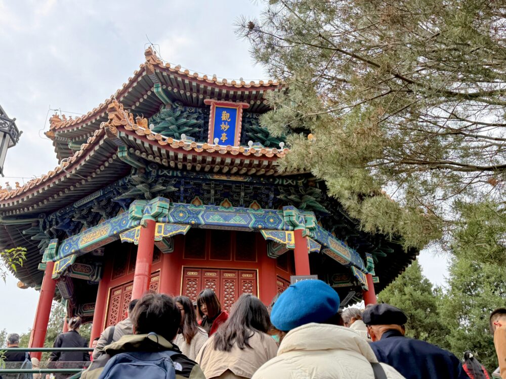景山公園の山頂にある「観妙亭」を下から見上げた写真。赤い柱と青・緑・金の極彩色の装飾が施された八角形の伝統的な中国建築で、多くの観光客が入口前に集まっている様子が写っている。