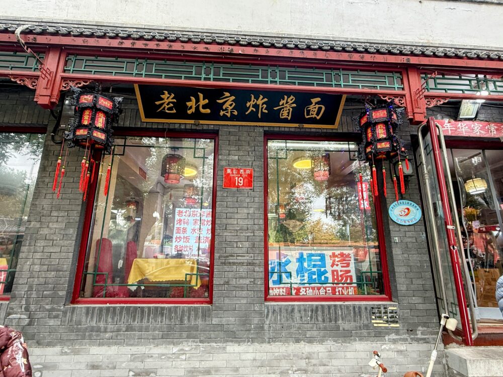 黒いレンガ造りの外観に赤いランタンが下がる「老北京炸醤麺」の店舗正面。大きな窓から店内のテーブル席が見える。