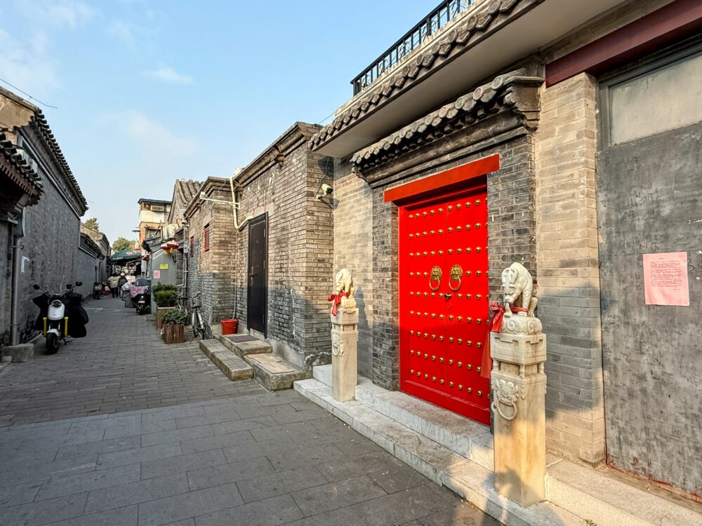 胡同の小道沿いに並ぶ伝統的な灰色煉瓦の住宅と、金色の飾りがついた赤い大門

