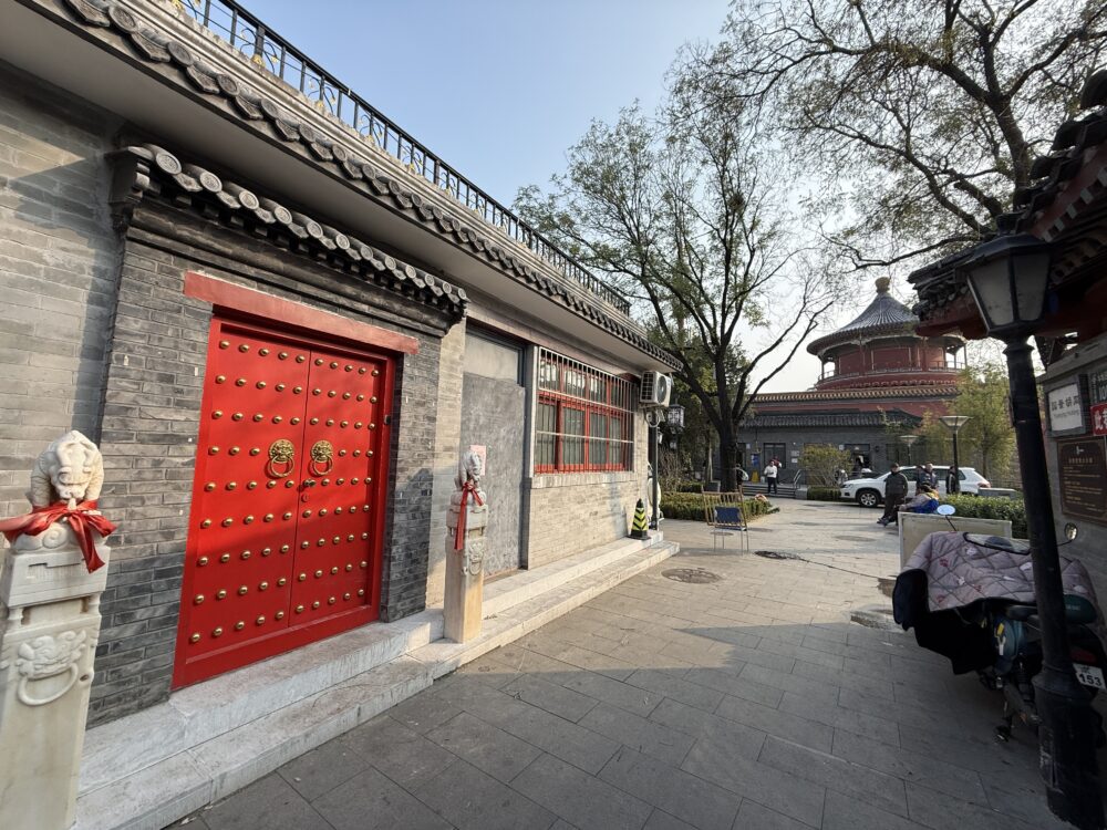 北京の胡同（フートン）を歩く途中に見つけた、赤い門が美しい伝統建築。奥には歴史ある円形の建物が見え、古都の雰囲気を感じる風景。
