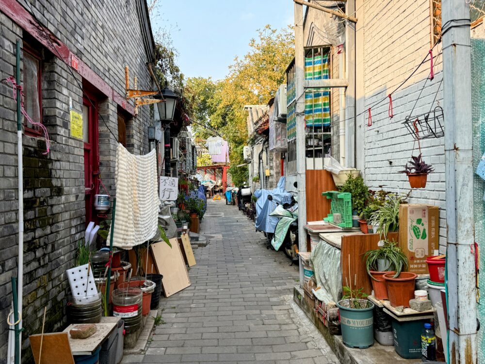 洗濯物や植木鉢、日用品が並ぶ細い胡同の小道。生活感のある家々が立ち並ぶ風景。