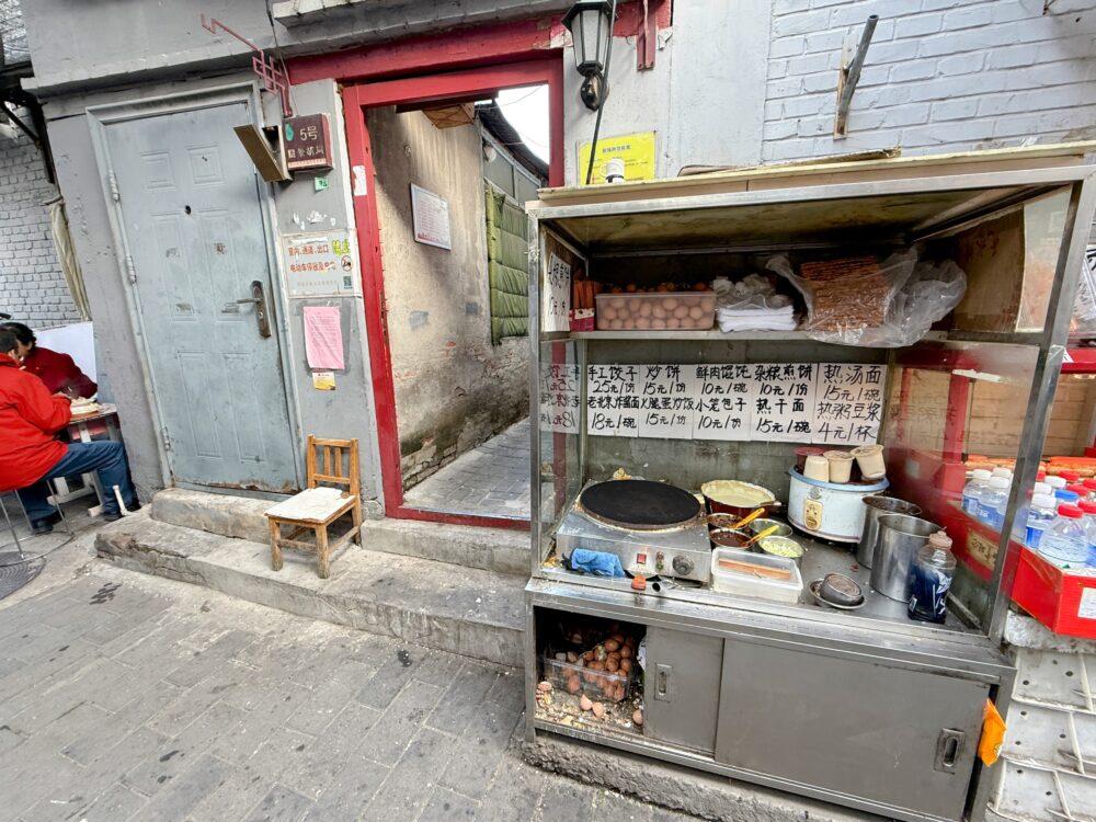 胡同の路地にある小さな食べ物屋台。鉄板や卵が並び、手工餃子などの手書きメニューが貼られている