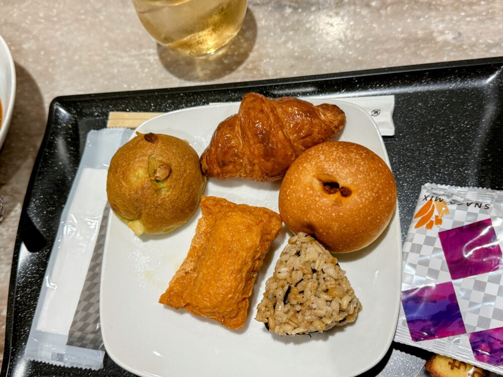 羽田空港ANAスイートラウンジで提供される軽食プレート。クロワッサン、いなり寿司、玄米おにぎり、ピスタチオパン、チーズパン、スナックミックスが並ぶ。